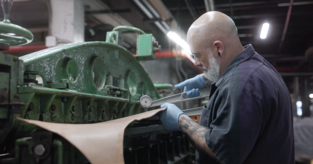 Worker feeding a strip of undyed leather into a green band-knife splitting machine at Horween, checking thickness with a dial gauge